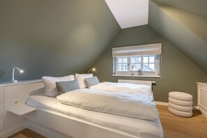Een slaapkamer met een schuine kap, een bed met wit beddengoed, een nachtkastje, een raam met uitzicht en een radiator.