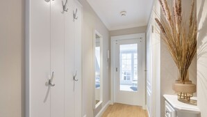 Un couloir avec des armoires blanches, un plancher en bois, une porte donnant sur un balcon et un vase contenant des plantes séchées, posé sur une console blanche.