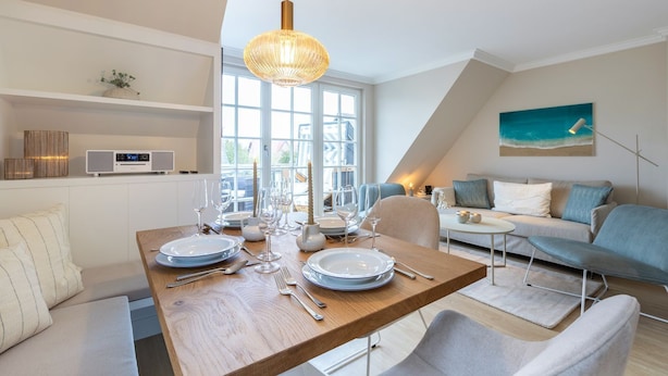 Un coin repas avec une table en bois dressée pour quatre personnes, un luminaire suspendu moderne et une vue sur l’extérieur grâce à de grandes fenêtres.