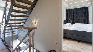 A modern hotel room with a glass staircase, a bed with dark bedding, and a bright ceiling light. - Willms Living, Long Beach House - Design-avantgarde. Viel Raum. Direkt an den Tinnumer Wiesen (Sylt)
