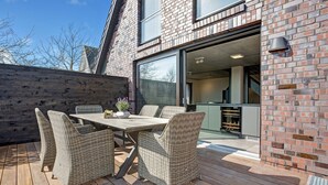 An outdoor patio with a wooden deck, wicker furniture, and a brick building with large windows. - Willms Living, Long Beach House - Design-avantgarde. Viel Raum. Direkt an den Tinnumer Wiesen (Sylt)