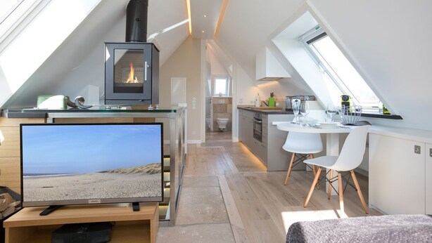 A modern loft bedroom with a large TV, a fireplace, and a dining area with a view of the sky. - Willms Living, Sky - Design [&] Stil Pur. Raumwunder für Individualisten an den Tinnumer Wiesen (Sylt-Ost)