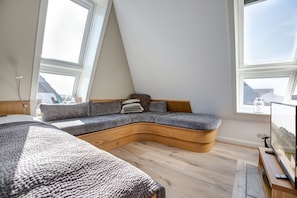 A loft bedroom with a curved wooden bed, a built-in seating area with cushions, and large windows offering a view of the sky. - Willms Living, Sky - Design [&] Stil Pur. Raumwunder für Individualisten an den Tinnumer Wiesen (Sylt-Ost)