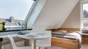 A high-ceilinged room with a dining table set for two, a cozy bed with a wooden headboard, and a large window offering a view of a building outside. - Willms Living, Sky - Design [&] Stil Pur. Raumwunder für Individualisten an den Tinnumer Wiesen (Sylt-Ost)
