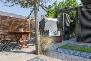 A patio area with a wooden table, a lounge chair, and a small garden. - Willms Living, Sky - Design [&] Stil Pur. Raumwunder für Individualisten an den Tinnumer Wiesen (Sylt-Ost)