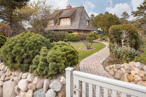 Uma casa com telhado de colmo, rodeada por um jardim bem cuidado com arbustos variados e um caminho de pedra que leva à entrada.
