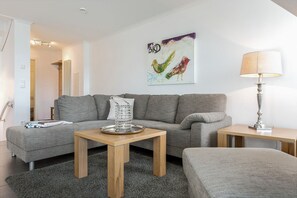 Een moderne woonkamer met een grijze bank, een salontafel en een lamp. Aan de muur hangt een schilderij en er is een deur naar een andere kamer.