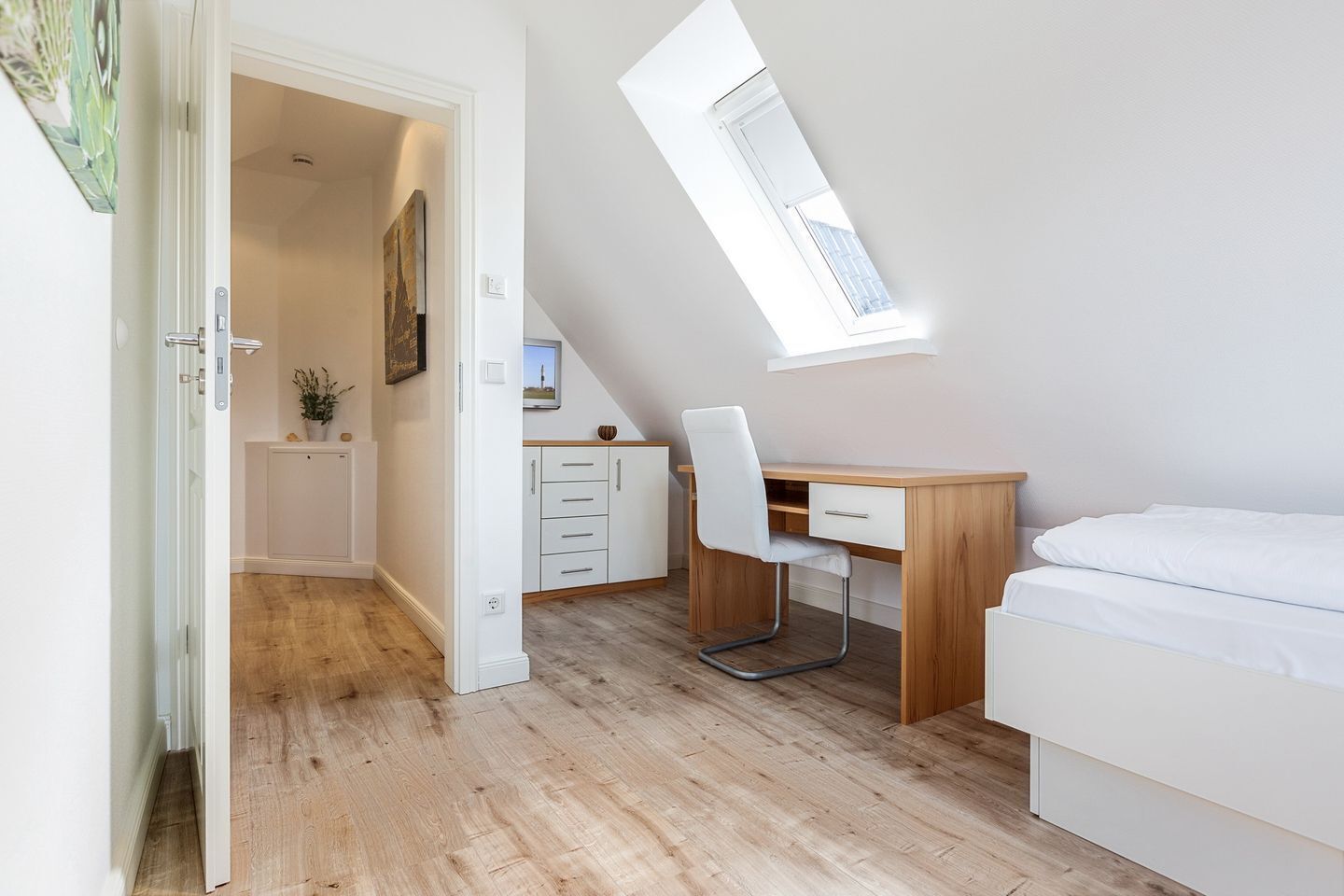 Ein Schlafzimmer mit schrägen Wänden, einem Oberlicht, einem Holztisch, einem weißen Stuhl und einem Bett.
