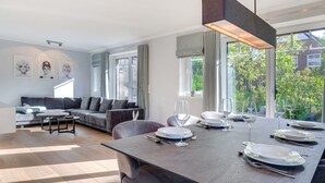 Un comedor moderno con una mesa de madera preparada para cuatro personas, un sofá gris y un amplio ventanal con vistas a zonas verdes.