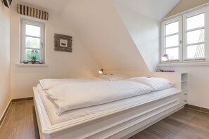 A bedroom with a bed, two windows, a bedside table, and a small shelf. - Oceanhouse, Lichtmeer, App. 7 - Strandnahe & Sogar Kleiner Meerblick. Überzeugender Luxus & a lot (Wenningstedt-Braderup (Sylt))
