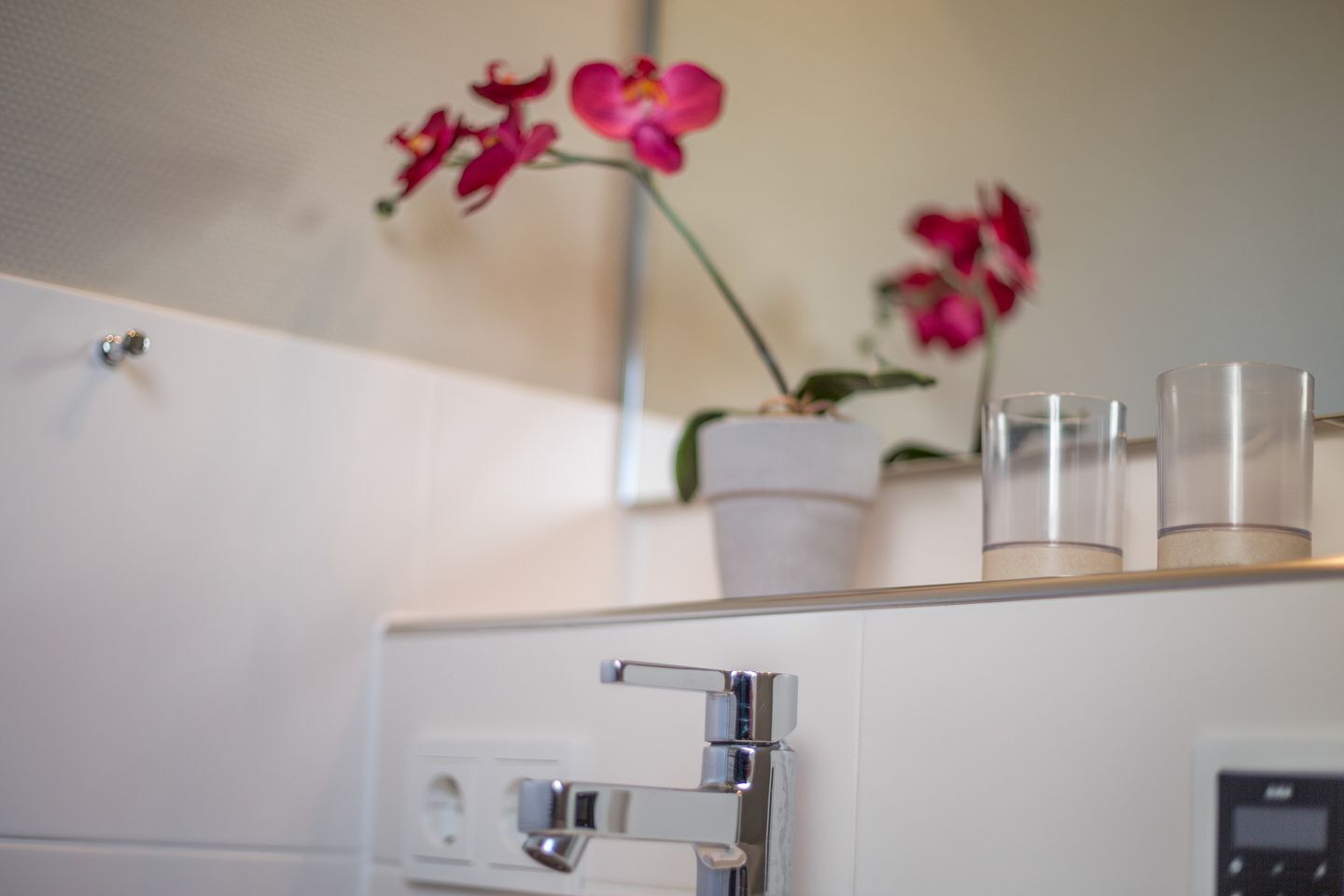 Ein weißes Schränkchen mit einer Metallgriff, eine Orchidee im Topf und zwei klare Gläser darauf.