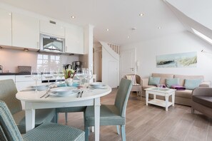 A modern kitchen with white cabinets and a built-in oven. There is a dining area with a round table and chairs, and a living room with a sofa and a coffee table. - Oceanhouse, Lichtmeer, App. 7 - Strandnahe & Sogar Kleiner Meerblick. Überzeugender Luxus & a lot (Wenningstedt-Braderup (Sylt))