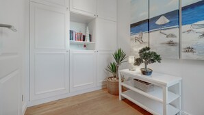Una stanza con un armadio bianco a muro, un tavolino consolle bianco con una lampada e un bonsai, e una parete con tre quadri raffiguranti una spiaggia.