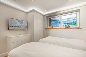 Un dormitorio con televisor, ventana con vistas al mar, estante de madera y cómoda blanca.