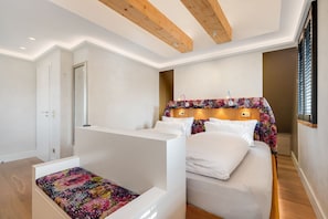 A bedroom with a bed, a small seating area, wooden beams on the ceiling, and a window with blinds.