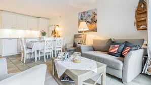 A modern living room with a grey sofa, a dining area with white chairs, and a kitchen with white cabinets.