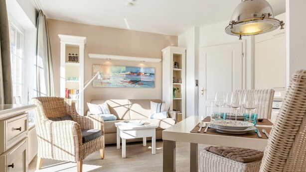 A well-lit room with a dining table set for two, a sofa, wicker chairs, and a painting on the wall. - Haus Am Dorfteich, App. 2 - 2-raum-appartement im Erd- und Untergeschoss mit ca. 40m² Nutzfläche (Wenningstedt-Braderup (Sylt))