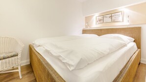 A neatly made bed with white linens, a wicker chair, and a framed picture on the wall. - Haus Am Dorfteich, App. 2 - 2-raum-appartement im Erd- und Untergeschoss mit ca. 40m² Nutzfläche (Wenningstedt-Braderup (Sylt))