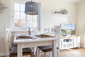 A dining area with a white table set for four, a television, and a window with a view of trees. - Haus Am Dorfteich, App. Élise - Idylle & Komfort Unmittelbar am Dorfteich (Wenningstedt-Braderup (Sylt))