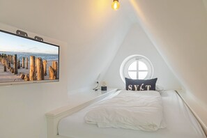 Ein Schlafzimmer mit großem Fenster, einem Bett mit weißer Bettwäsche, einem Kopfkissen mit dem Aufdruck 'SYLT' und einem Bild von Holzpfählen an einem Strand.