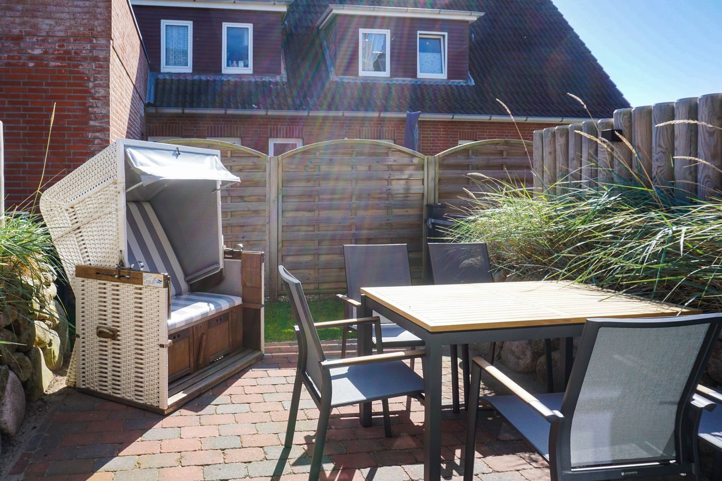 Een terras buiten met een tafel en stoelen, een zithoek met kussens en een houten omheining.