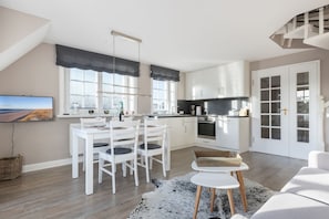 A modern kitchen with white cabinets, a dining area with white chairs, and a large window with blinds. - Windlicht - 3- Raum Appartement im Og/dg mit ca. 54qm Nutzfläche in Munkmarsch (Sylt-Ost)