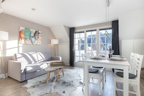 A modern living room with a grey sofa, a dining table set for four, and a large window with curtains. - Windlicht - 3- Raum Appartement im Og/dg mit ca. 54qm Nutzfläche in Munkmarsch (Sylt-Ost)