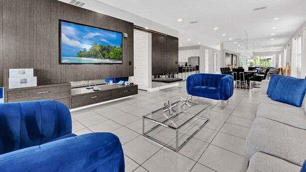 A modern living room with a large flat-screen TV, blue sofas, a glass coffee table, and a kitchen area in the background. - 8BR Modern Villa • Games&Movie Room • Near Disney (Kissimmee)