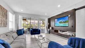 A modern living room with a large flat-screen TV, a sofa, a coffee table, and a glass door leading to a balcony. - 8BR Modern Villa • Games&Movie Room • Near Disney (Kissimmee)