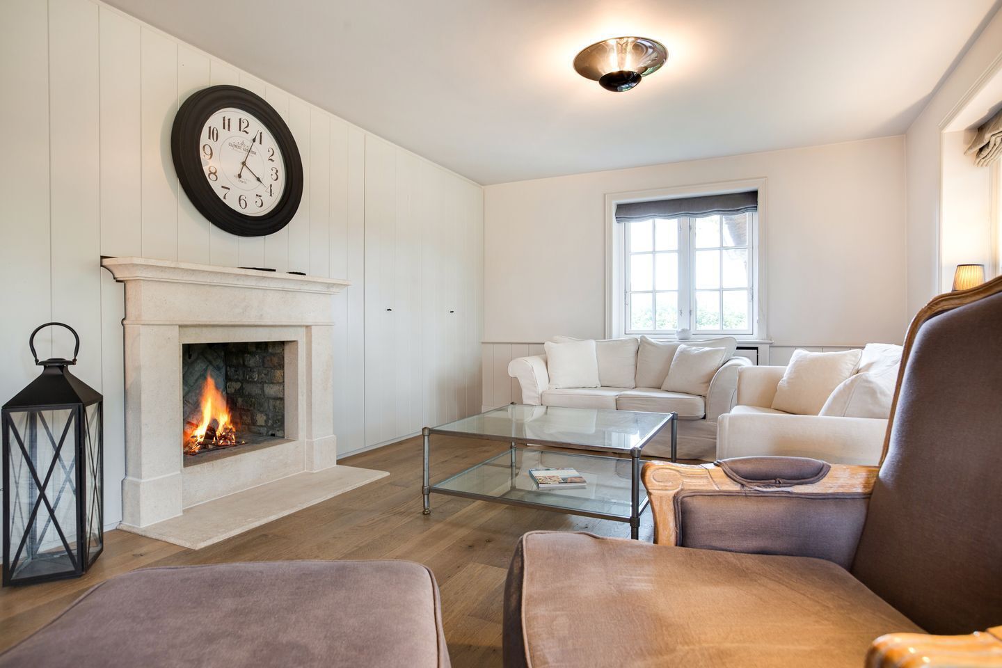 Un salon avec une cheminée, une horloge murale ronde, une table basse en verre et un ensemble de canapés blancs.