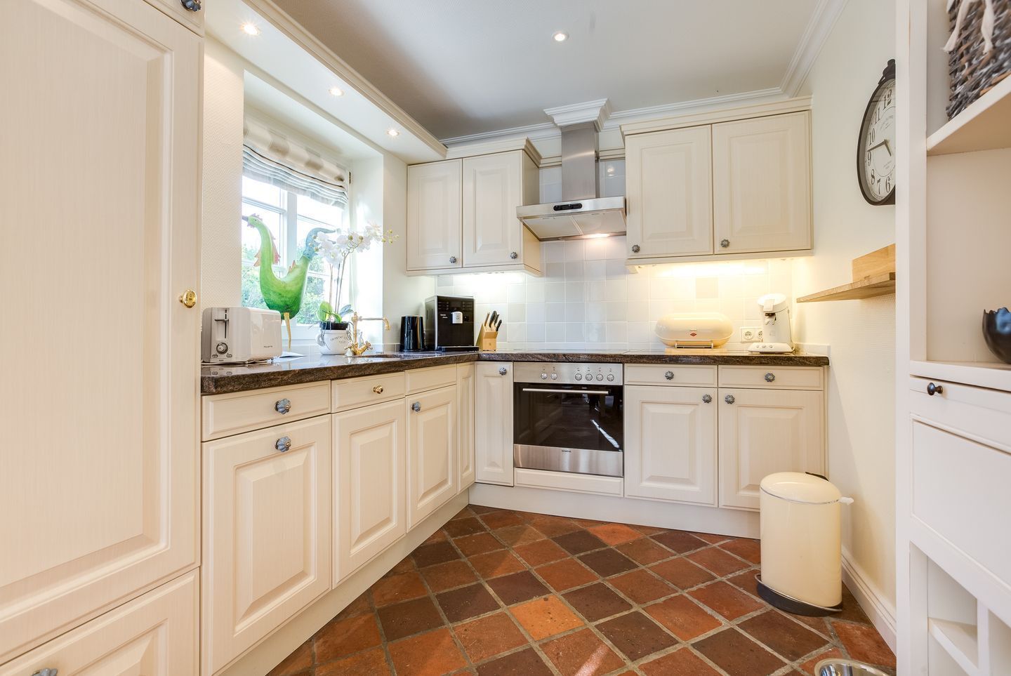 Una cocina con armarios blancos, horno de acero inoxidable, cafetera y ventana con planta decorativa.