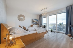 Et moderne soveværelse med en stor seng, et natbord med lampe, et fjernsyn og udsigt over byen gennem store vinduer.