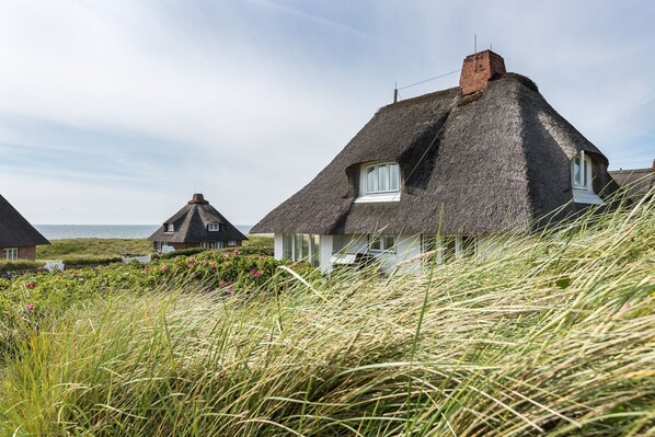 Ein Haus mit einem Reetdach und weißer Fassade, umgeben von hohem Gras unter blauem Himmel.