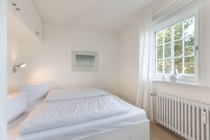 Une chambre à coucher blanche avec un lit, une lampe fixée au mur, un tableau encadré et une fenêtre avec des rideaux.