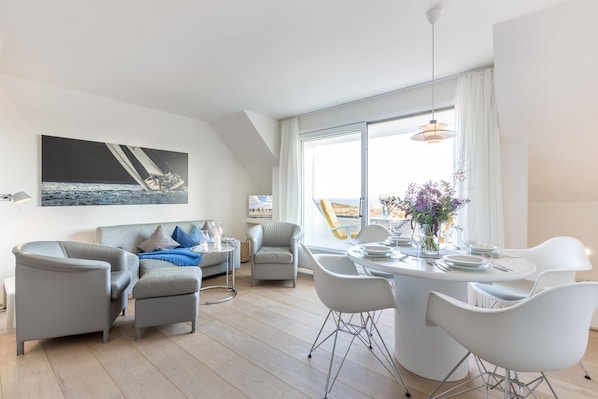 Un salon moderne avec un canapé, des fauteuils, une table à manger pour quatre personnes et une grande fenêtre donnant sur la mer.