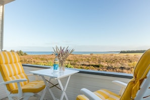 Un balcon avec des chaises à rayures jaunes, une petite table avec une bouteille et des verres, et une vue sur un paysage côtier.