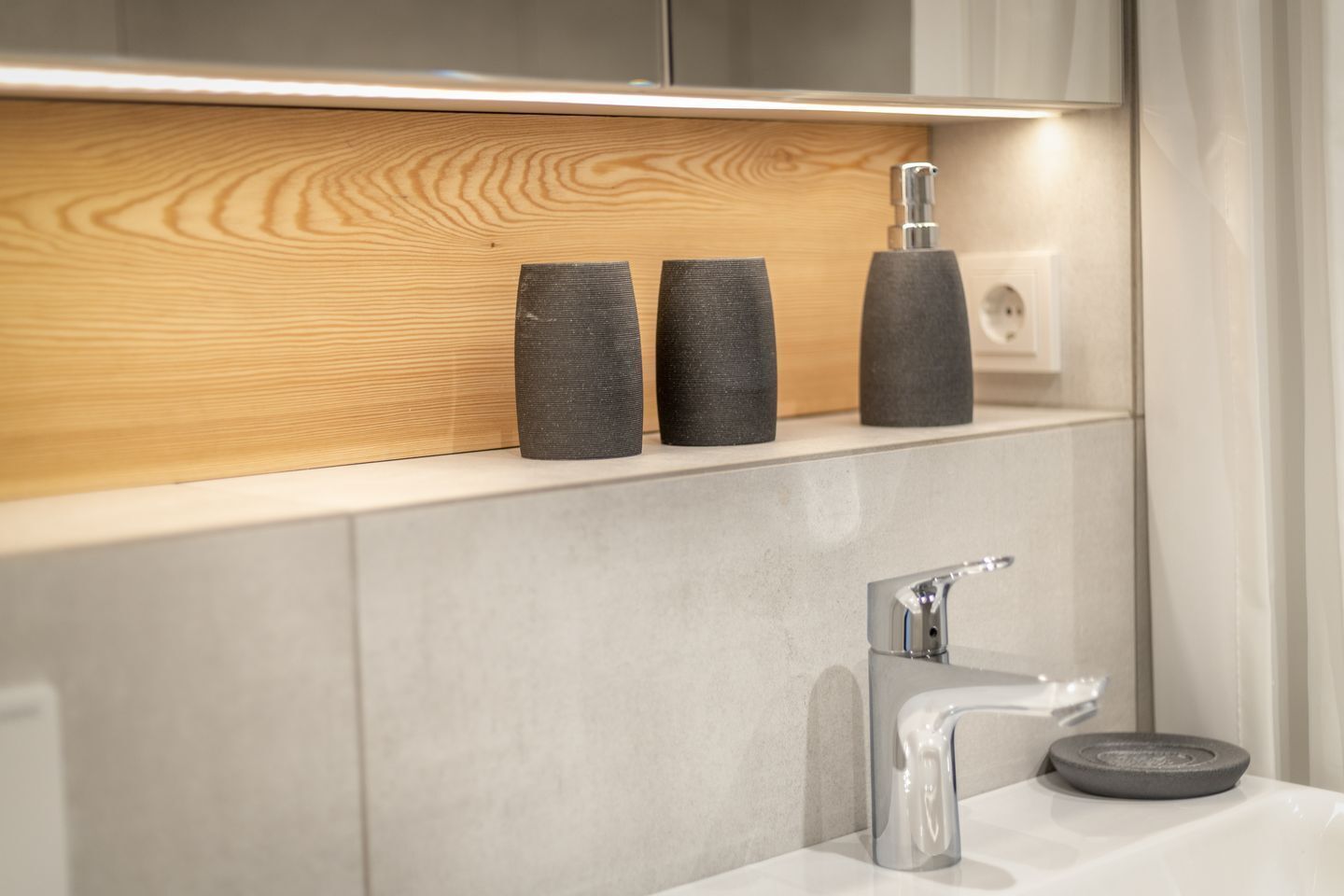 Ein modernes Badezimmer mit Ablagefach aus Holz, drei schwarzen Behältern, einem Chromarmaturen und einer Seifenschale.
