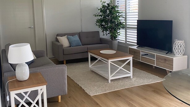 A modern living room with a grey sofa, two white coffee tables, a TV, and a potted plant. - Rural Setting - Close to shops, Townsville Hospital and Riverway (Gumlow)
