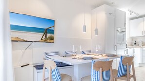 A modern kitchen with white cabinets, a dining area with striped chairs and a table set for four, and a large beach-themed painting on the wall. - Ringelgans - Urlaub im Herzen von Westerland, das Erleben Sie in dem Appartement Ringelgans (Sylt)