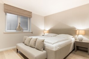 A bedroom with a bed, bedside tables, a bench, a window with a curtain, and a lamp. - Düün Hüs, App.2 - Ferienwohnung "düün Hüs, App. 2" in Westerland, Sylt (Sylt)