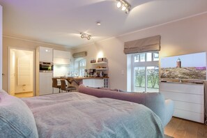 A bedroom with a large bed, a desk, and a TV. There is a kitchen area with a stove and a window. - Inselsuite - Beste Lage. Beste Ausstattung. Bester Urlaub (Sylt)