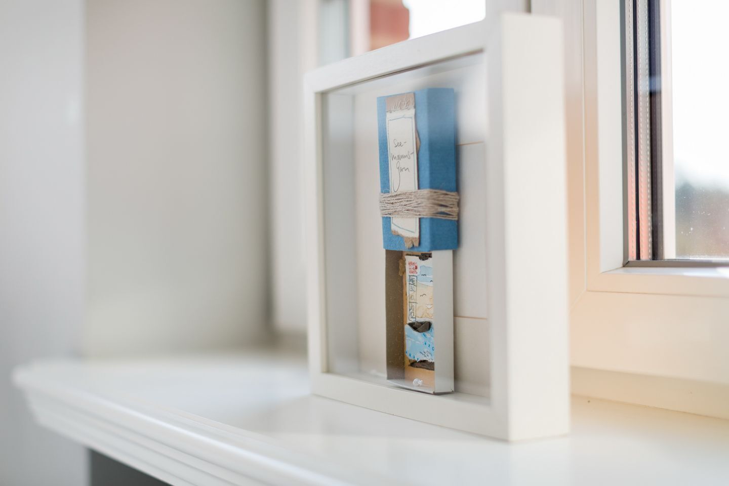 A framed picture with a handwritten note and a blue card on a white window ledge.