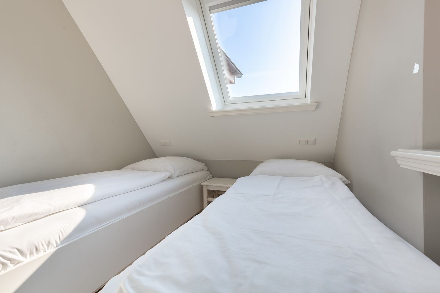 Une petite chambre avec un seul lit, deux lits simples, une lucarne et une décoration aux tons blancs.