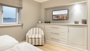 Une chambre à coucher avec un lit, un fauteuil à rayures, un téléviseur posé sur une étagère et une vue sur la mer par la fenêtre.
