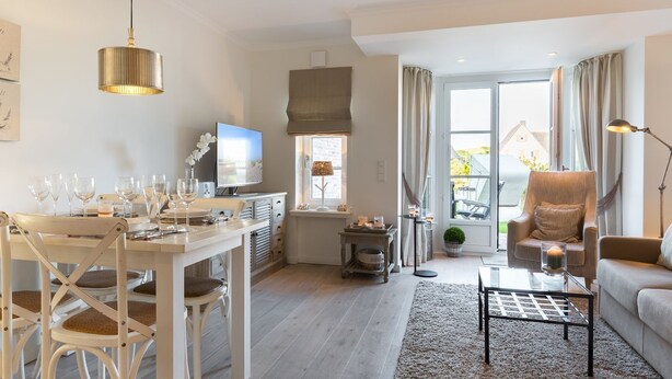 A modern living room with a dining area, a television, and a large window with curtains. - Dünen & Meer, App. 2 Dünen-nest - In Strandnaher und Dennoch Zentraler Lage Gelegenes 3-raum (Sylt)