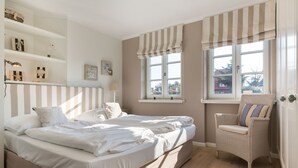 A bedroom with a large bed, two bedside tables, a chair, and a window with curtains. - Dünen & Meer, App. 2 Dünen-nest - In Strandnaher und Dennoch Zentraler Lage Gelegenes 3-raum (Sylt)