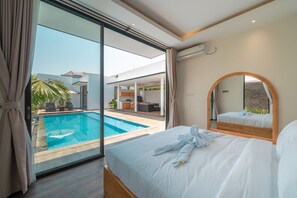 A modern hotel room with a large bed, a bathroom with a mirror, and a view of an outdoor swimming pool and patio area. - Villa Selikur 1  (Seminyak)