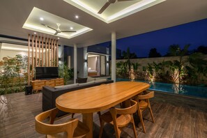 A modern outdoor living area with a wooden table and chairs, a TV, and a comfortable seating area. - Villa Selikur 1  (Seminyak)