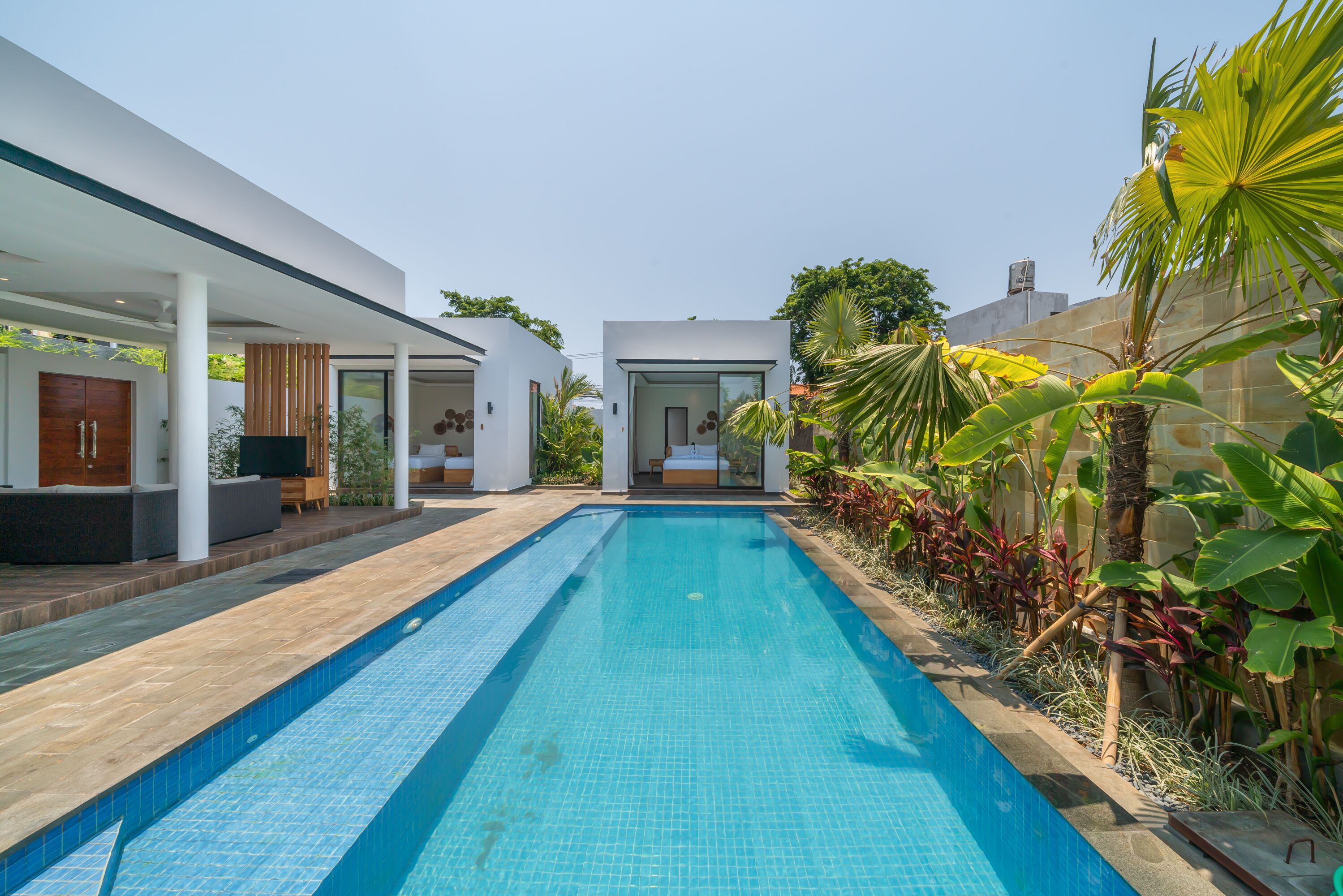 A modern pool area with a clear blue pool, surrounded by lush greenery and a contemporary building with large windows and a flat roof.