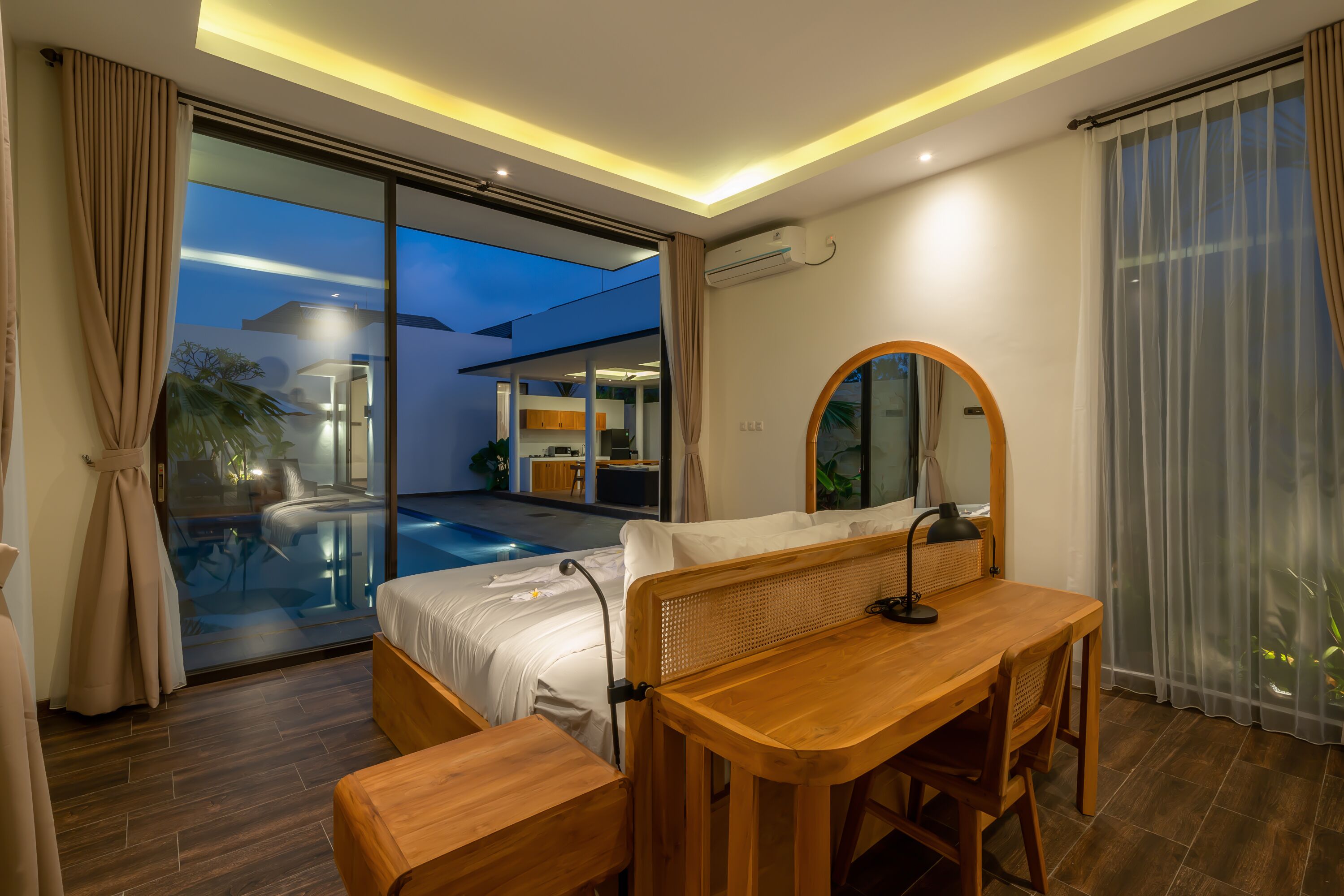 Quarto de hotel moderno com uma cama grande, uma escrivaninha de madeira, um espelho redondo e uma porta de vidro que dá para uma área de piscina.
